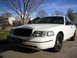 2000 Ford Crown Victoria - A white 2000 Ford Crown Victoria is parked in a residential driveway under a clear blue sky.