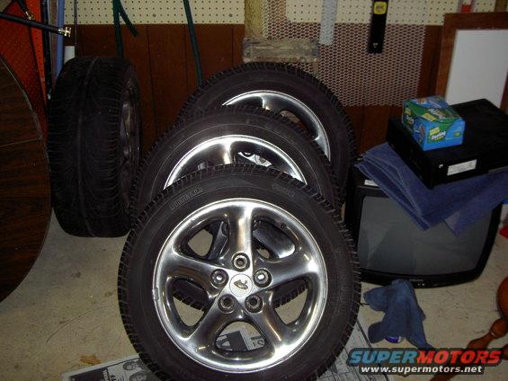 Four shiny alloy wheels with tires stacked in a garage, next to an old TV