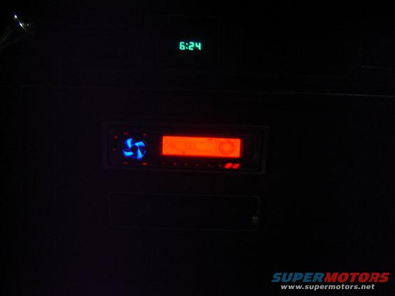 Car stereo with orange display and blue fan, above it a green digital clock reads 6:24