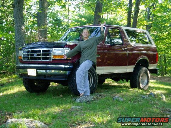 Maroon and white Ford Bronco with a lifted suspension and off-road tires, parked in a wooded area