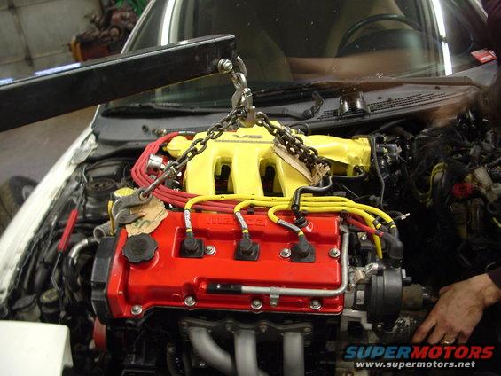 Engine with red valve cover and yellow intake manifold being lifted by a chain hoist