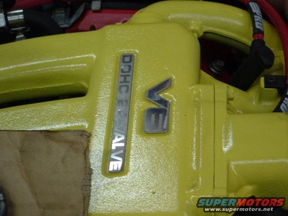Close-up of a yellow engine component with "DOHC 24 VALVE" and "V6" emblems. Some brown cardboard is visible on the left