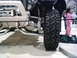 1986 Ford Bronco - Close-up view of an off-road vehicle's front suspension, large tire, and winch, with a child in the background.