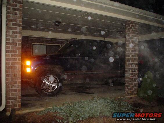 Side view of a dark pickup truck parked under a carport at night. The truck has its front turn signal illuminated