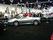 1986 Ford Bronco - A silver convertible sports car is shown at an auto show with people in the background.