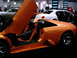 1986 Ford Bronco - An orange sports car is displayed with its doors open, parked next to a white sedan.