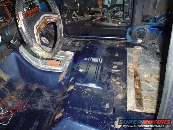Interior floor of a vehicle with exposed metal, rust, and wiring. Steering wheel visible