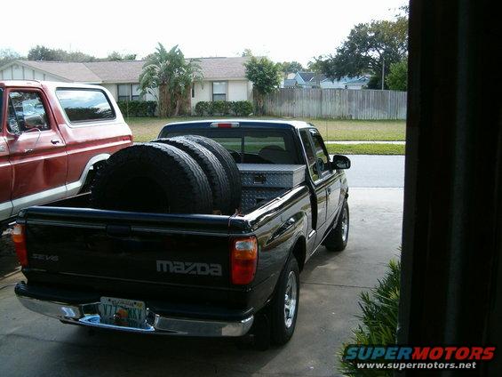 Black Mazda pickup truck rear view, carrying three tires and a toolbox