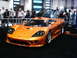 1986 Ford Bronco - An orange sports car is displayed indoors on a black carpeted floor.