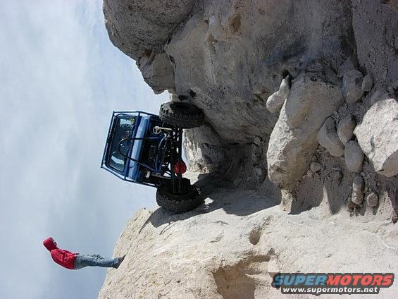 Blue off-road vehicle precariously balanced on a rocky cliff face with a person in a red jacket nearby