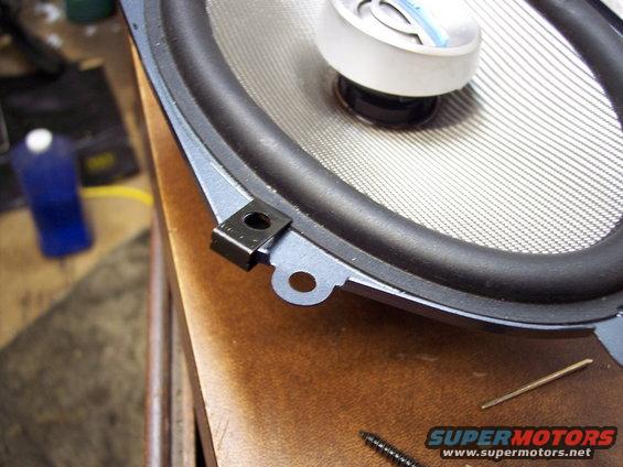 Car speaker with a metal grille and black rubber surround, resting on a wooden surface next to a screw