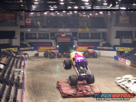 Monster truck with purple and yellow paint jumps over a crushed car in an arena