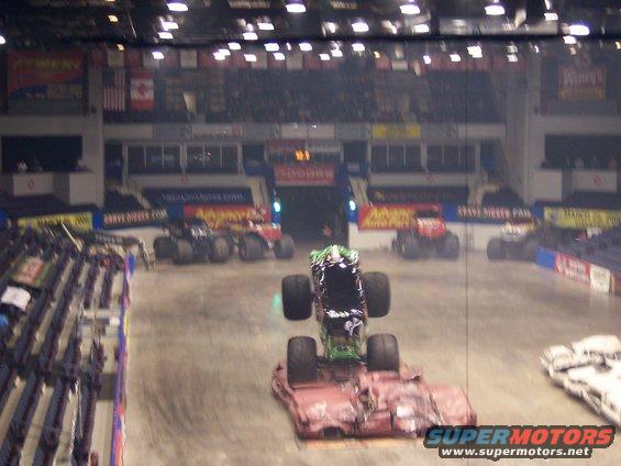 Monster truck on top of a crushed car in an arena with other trucks in the background