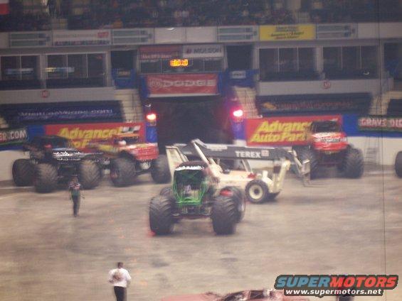 Monster trucks gathered in an arena, with a Terex crane in the center