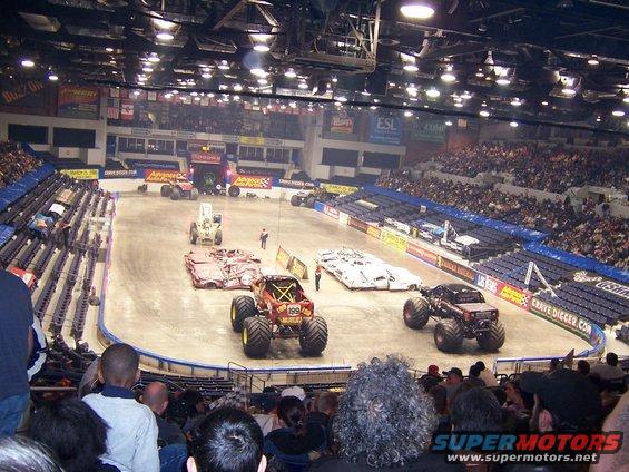 Monster trucks are lined up in an arena with spectators in the stands