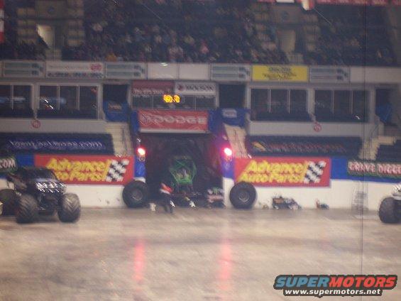 Monster truck with large tires in an arena, with other vehicles and spectators in the background