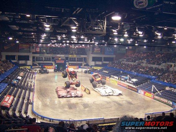 Monster trucks perform stunts in an arena, crushing cars below
