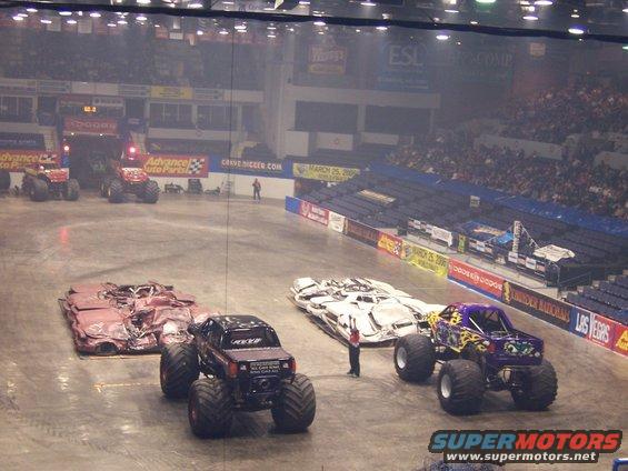 Monster trucks lined up on a concrete floor inside an arena with spectators in the stands
