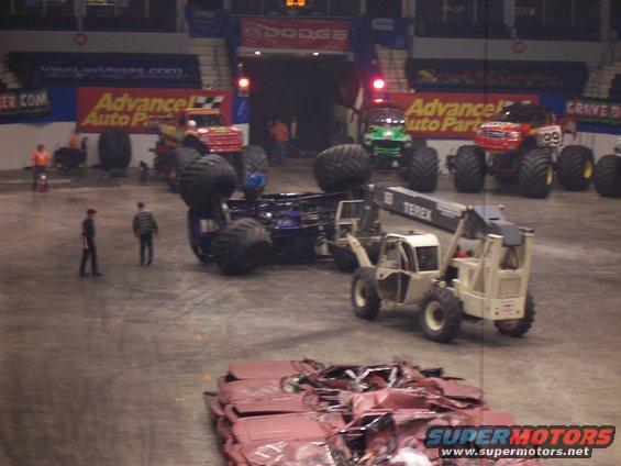 Monster truck overturned on its side, with a forklift nearby and crushed cars in the foreground