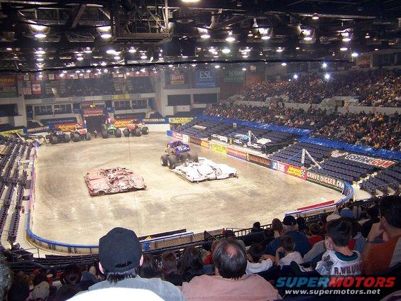 Monster trucks lined up in an arena with crushed cars on the floor and spectators in the stands