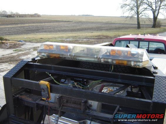 Light bar and wiring on a tow truck bed, with a red pickup truck visible behind it