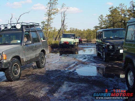 Several off-road vehicles navigate a muddy trail with trees and puddles