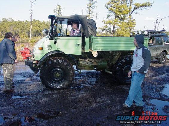 Unimog truck in muddy terrain with three people and a dark SUV in the background. Watermark reads 'SUPERMOTORS.NET'