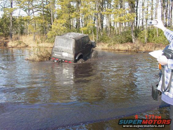 Suzuki Samurai partially submerged in muddy water, with trees in the background. A person holds a camera and paper on the right