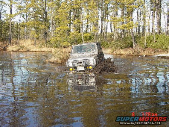 Suzuki Samurai driving through a muddy water crossing with trees in the background