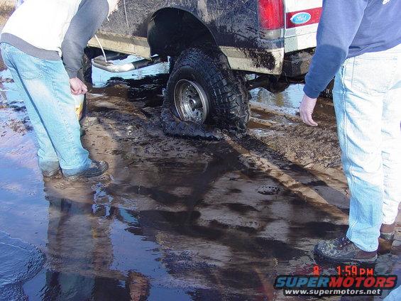 Mud-splattered truck rear with a flat tire stuck in mud, two people nearby