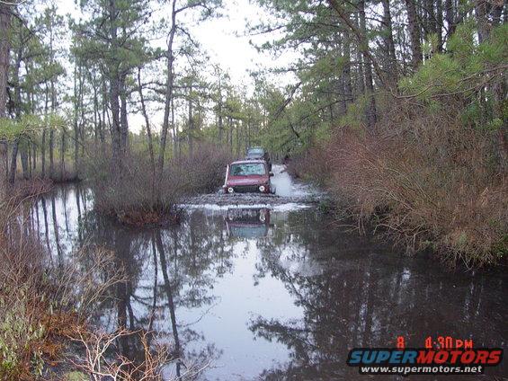 Red SUV driving through a muddy water crossing in a wooded area, with another vehicle behind it