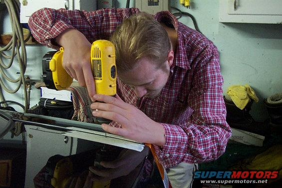 Man in red plaid shirt drills into shiny black car panel with yellow drill