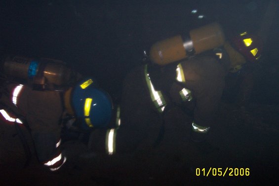 Two firefighters wearing yellow helmets and tan gear with reflective stripes, one with a yellow oxygen tank
