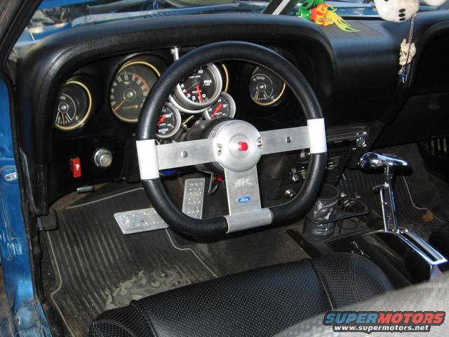 Car interior showing aftermarket steering wheel, gauges, and shifter. Ford logo visible on steering column