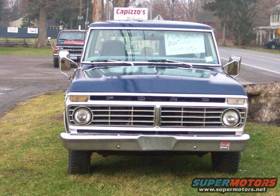 Front view of a blue Ford pickup truck with a 'For Sale' sign in the window. Another red truck is parked behind it