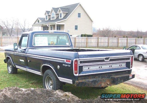 Dark blue Ford pickup truck, rear view. Has a metal bumper and a sign in the back window. Another car is parked nearby