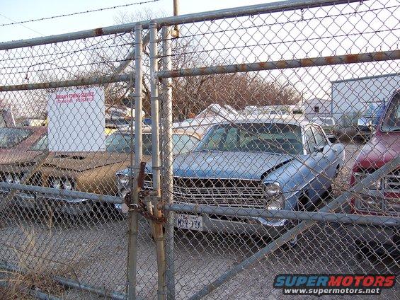 Blue sedan with chrome grille and dented hood, seen through a chain-link fence