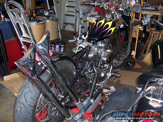 Custom motorcycle with flame decals on the tank, chrome exhaust pipes, and a tall sissy bar, parked indoors