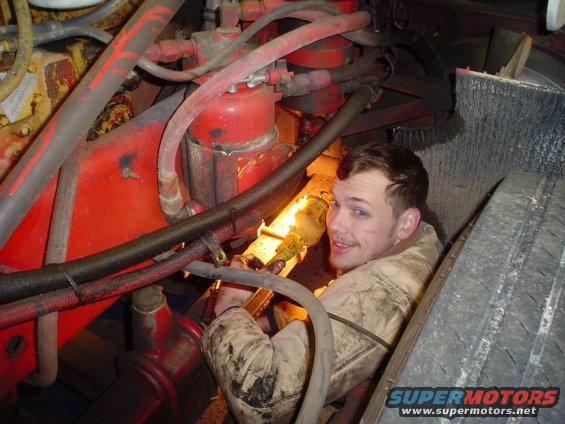 Mechanic in dirty overalls working on a red engine block with hoses and pipes; tire visible on right