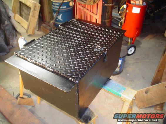 Black metal toolbox with diamond plate lid, resting on wooden supports. Red sandblaster in background