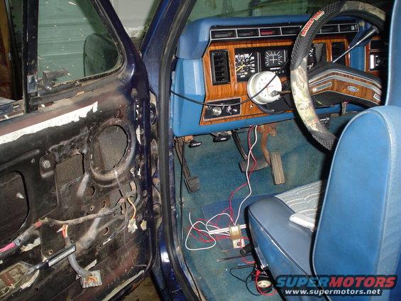 Interior of a Ford Bronco showing exposed door wiring, dashboard with wood grain trim, and blue seats. Aftermarket gauge mounted on steering column