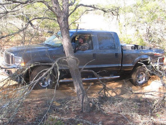 Dark gray Ford F-250 Super Duty pickup truck with a diamond plate toolbox in the bed, driving off-road through brush. Driver wears camouflage
