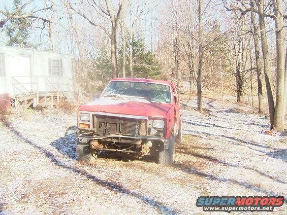 red pickup truck with missing grille and headlights, parked in a snowy, wooded area next to a trailer