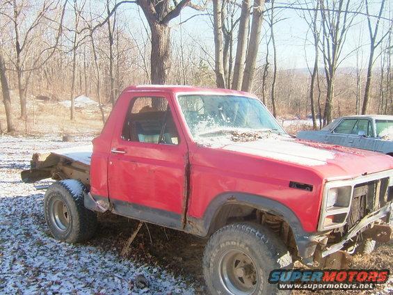 Red chopped-top pickup truck with flatbed, 3/4 front view. Another vehicle is visible behind it