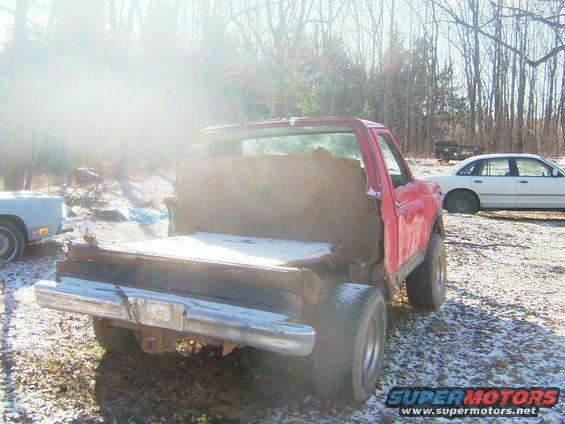 Red pickup truck with a custom wooden bed, seen from the rear. Another white sedan is parked nearby