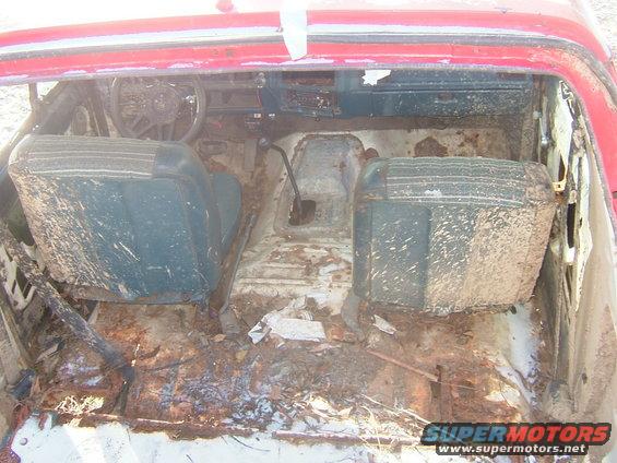 Interior of a heavily rusted and mud-covered red vehicle, showing front seats, dashboard, and floor