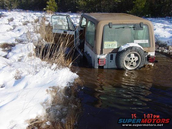 Brown and white Jeep Wrangler stuck in muddy water with snow on the bank. License plate SXW-45H is visible