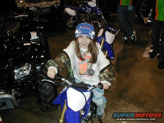 Child sits on a blue dirt bike. Black ATV and other motorcycles are visible in the background