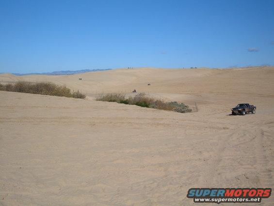 Dark colored pickup truck driving on a sandy dune under a clear blue sky. Other ATVs are visible in the distance