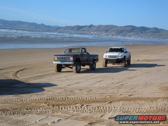 Brown pickup truck and white pickup truck on a sandy beach with ocean and mountains in the background